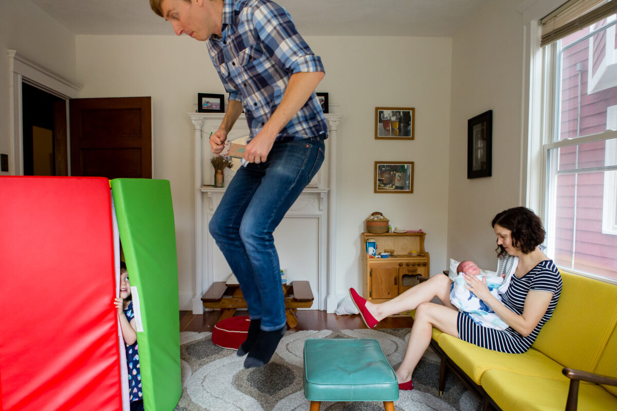Mom sits with her newborn on the couch while Dad plays with their 4-year-old daughter in Somerville, MA, by Emily Sterne Photography