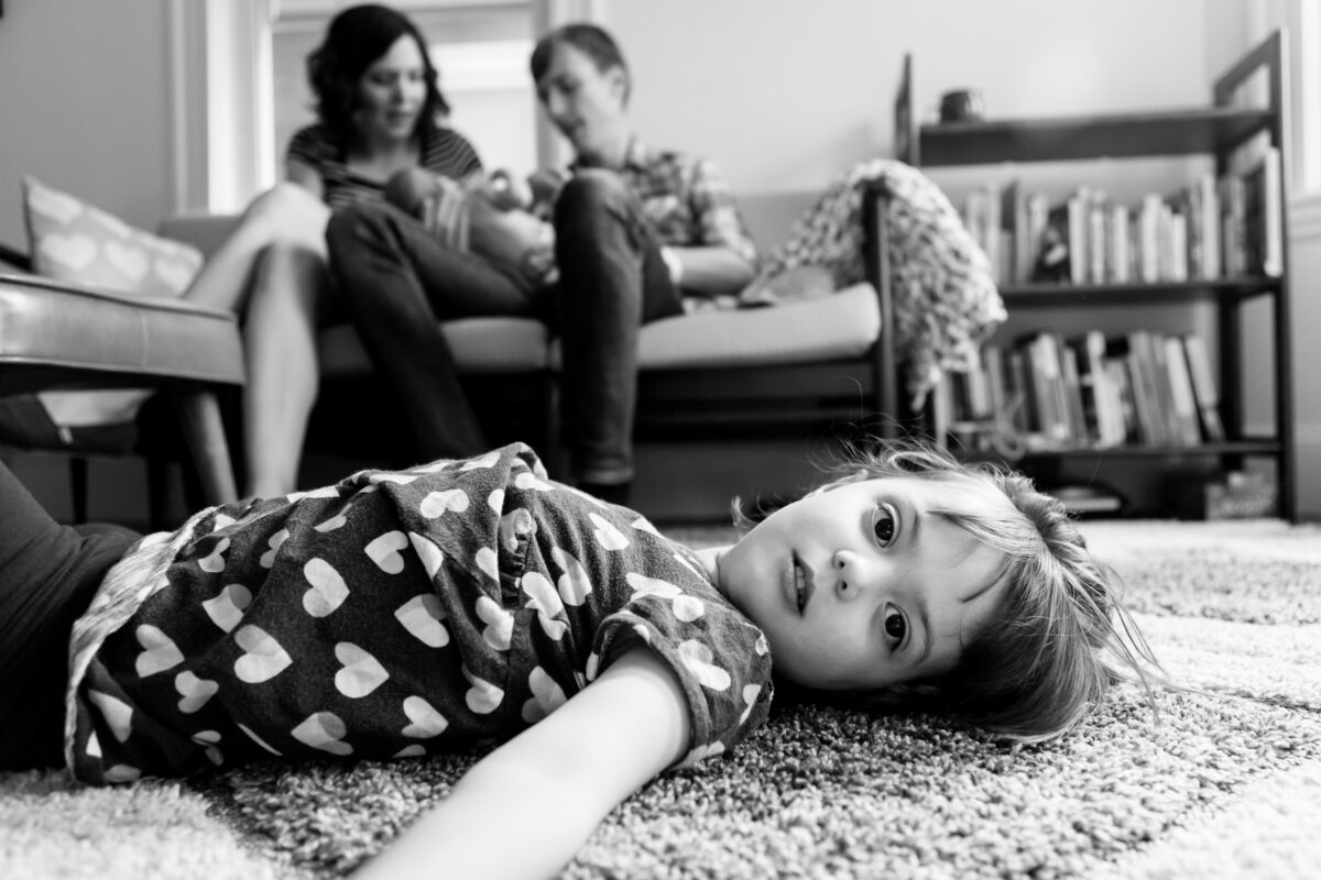 child lies on the carpet while mom and dad hold baby sibling on the couch