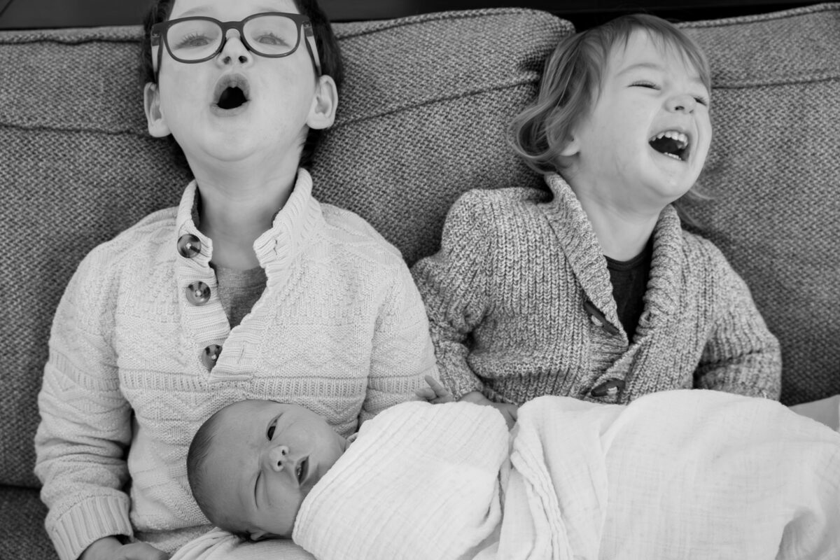 brothers make funny faces while balancing newborn sister on their laps during an in home session in Arlington, MA