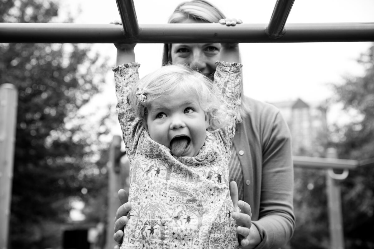 Young girl hangs from the monkey bars with help from mom and a silly face