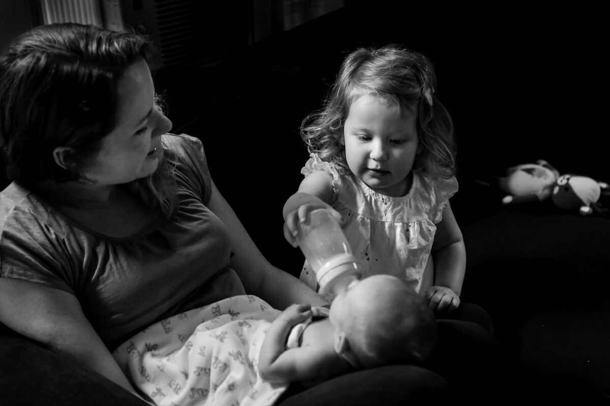 mom smiles at her toddler while they feed baby sister together on the couch