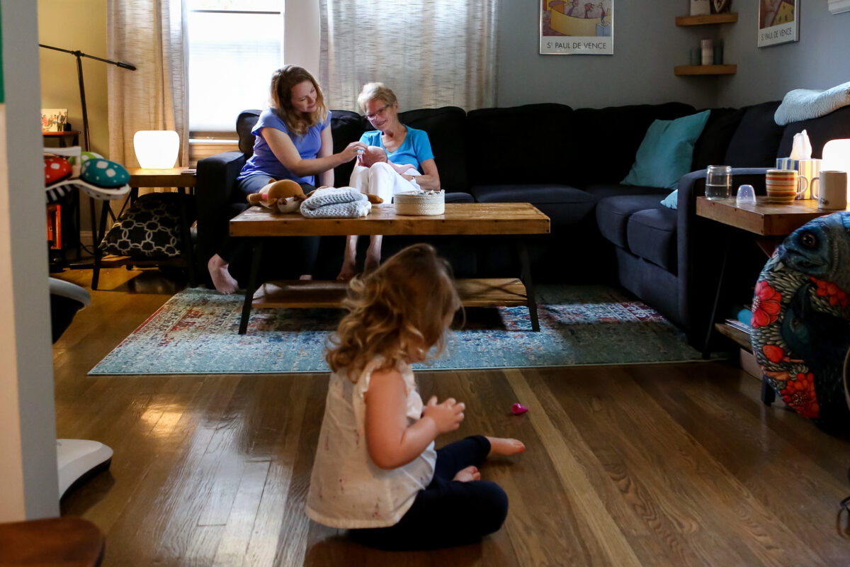 toddler plays on the floor as mom and grandma coo at baby sister in the background