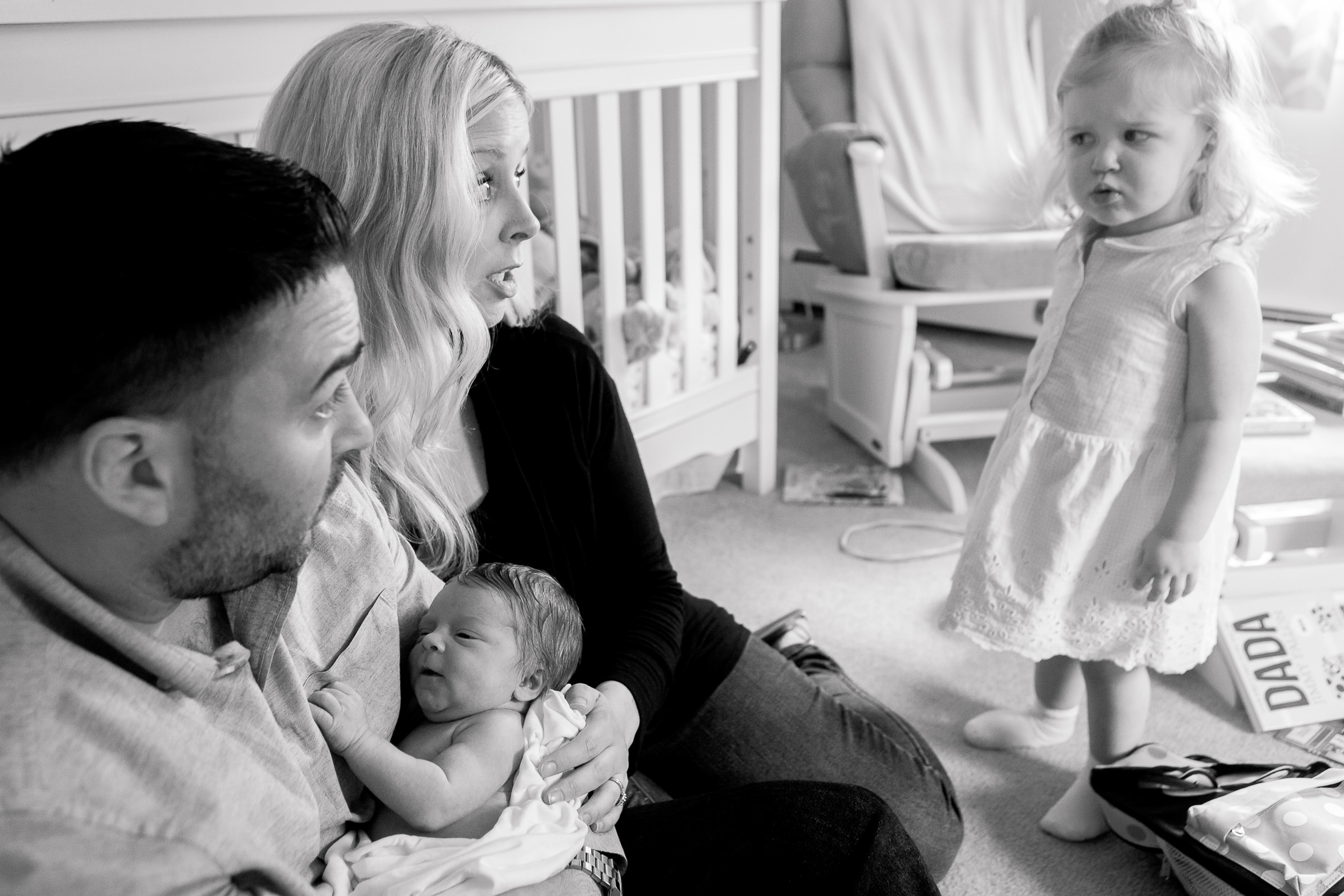 toddler gives mom and dad a skeptical look while they hold her newborn sibling